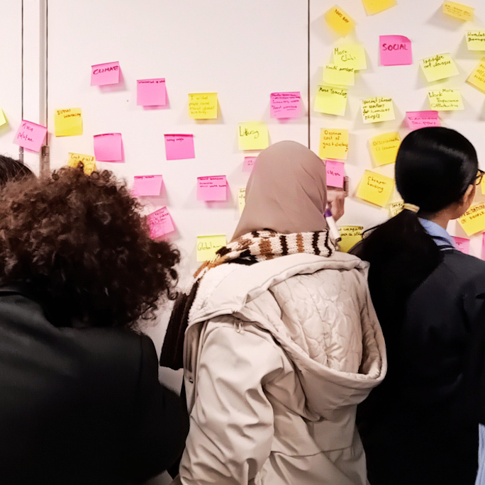 A group of teenagers from the Nova Youth Voice Group stand in front of a wall containing numerous post-it notes with ideas written on it, the group is voting which causes they should support