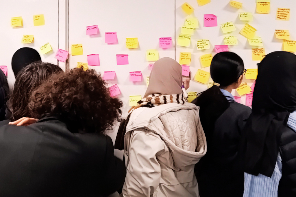 A group of teenagers from the Nova Youth Voice Group stand in front of a wall containing numerous post-it notes with ideas written on it, the group is voting which causes they should support